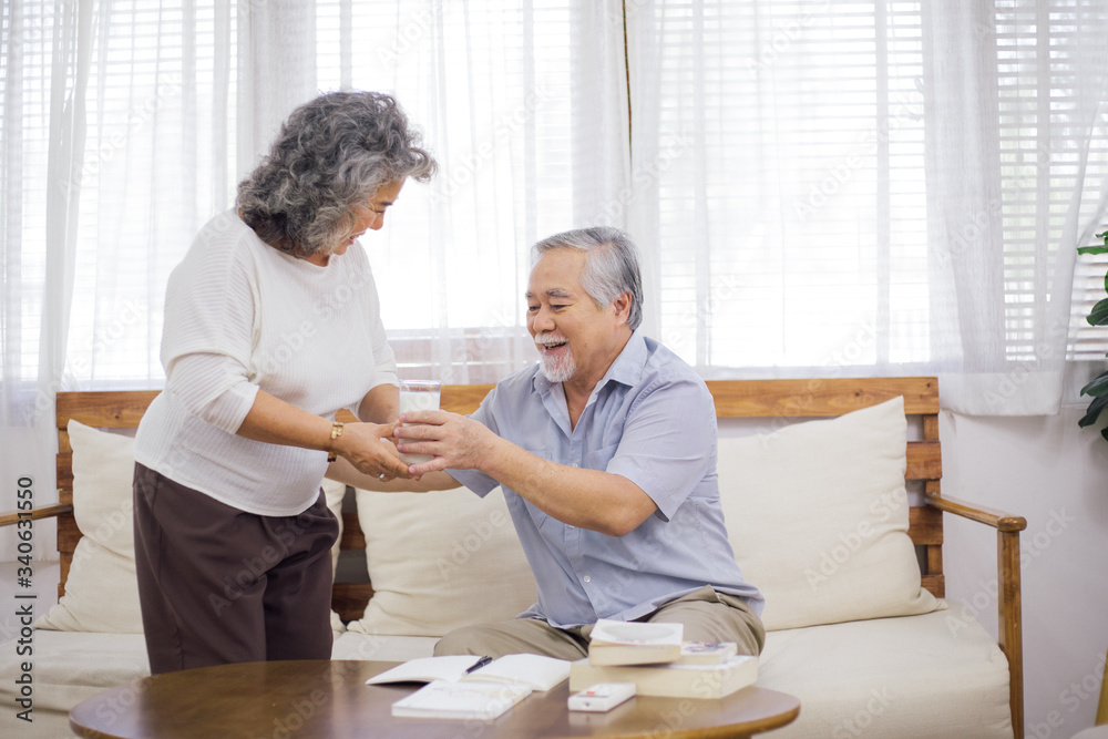 senior woman brink glass of milk to older husband in living room while he working or reading. self-quarantine from Coronavirus outbreak, love and take care family, healthy lifestyle insurance concept