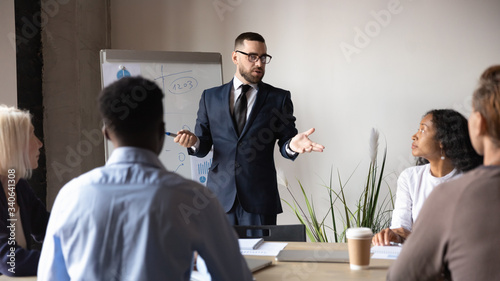 Concentrated male director in glasses lead meeting in office present business project on whiteboard, serious businessman make flip chart presentation for diverse multicultural colleagues or partners
