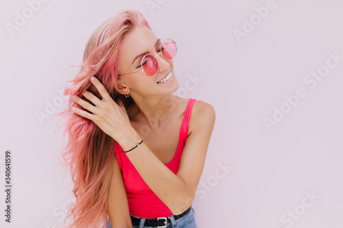 Charming girl wears earrings and bracelet touching her pink hair. Long-haired blissful woman posing with eyes closed in studio with white interior.