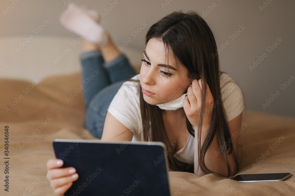 Young woman take off mask and use tablet on the bed. Female watch online video during quarantine. Student Study at home