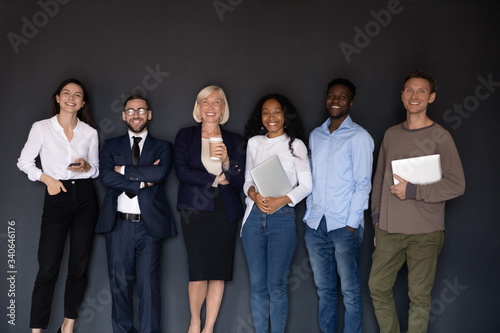 Group picture of smiling multiethnic diverse businesspeople stand posing near black wall look at camera together, happy multicultural employees make team photo at workplace, unity, teamwork concept