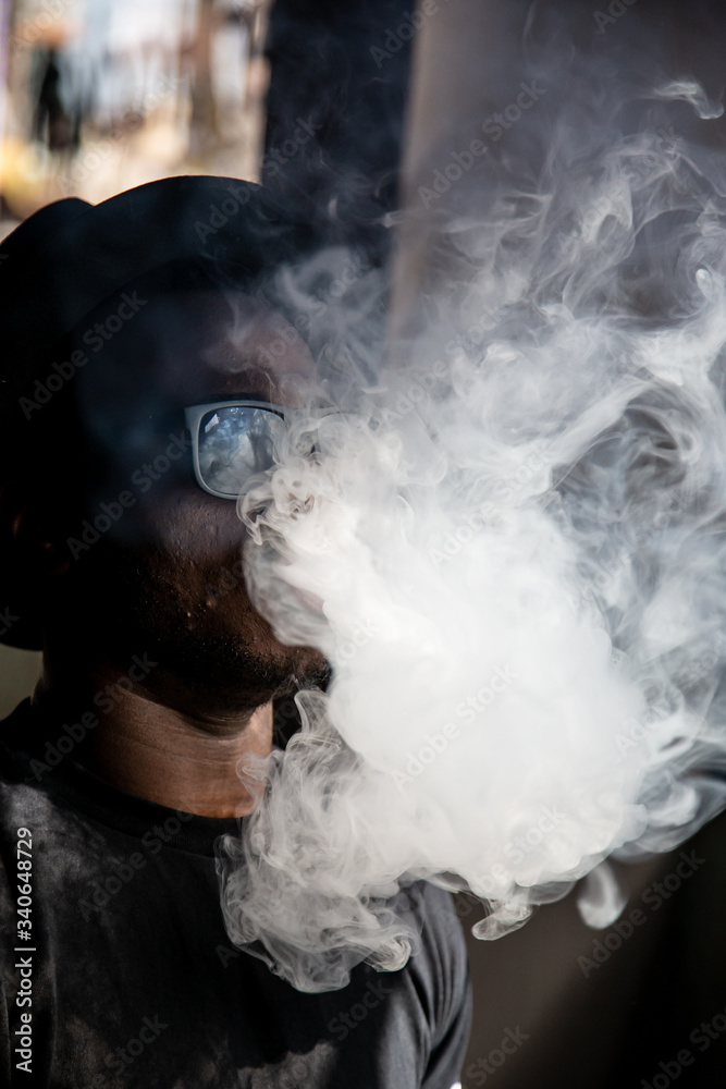 Vertical photo of a black young man with sunglasses and a vintage hat pulling smoke out