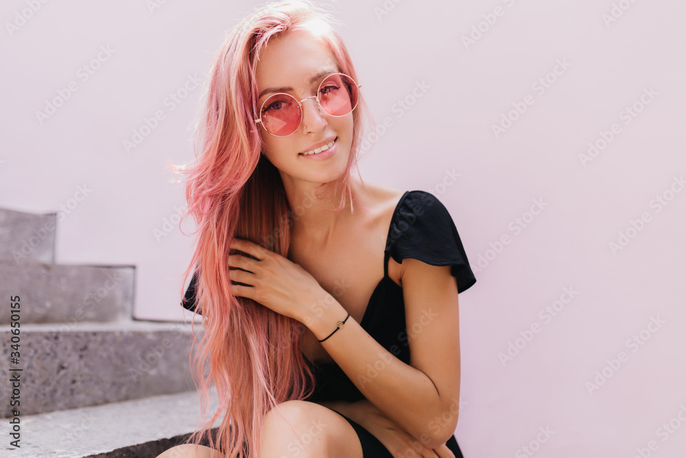 Fototapeta premium Wonderful caucasian lady sitting on stone stairs and touching her pink hair. Outdoor photo of lovely female model with tanned skin wears black attire.