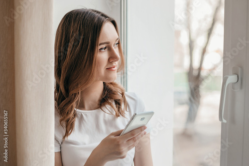 A young girl sits on a windowsill, looks at the window, holds a mobile phone and reads a message, searches for online remote work, coronavirus, quarantine, isolation, safe work