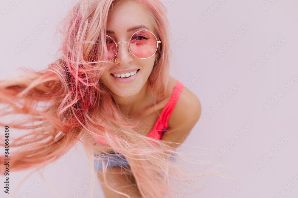 Close-up portrait of carefree beautiful girl playfully posing in studio. Excited lady in pink tank-top and sunglasses laughing on light background.