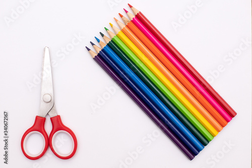 pencils of red, orange, yellow, green, azure, blue, and purple lie vertically on a white background in the shape of a rainbow next to a small pair of red scissors