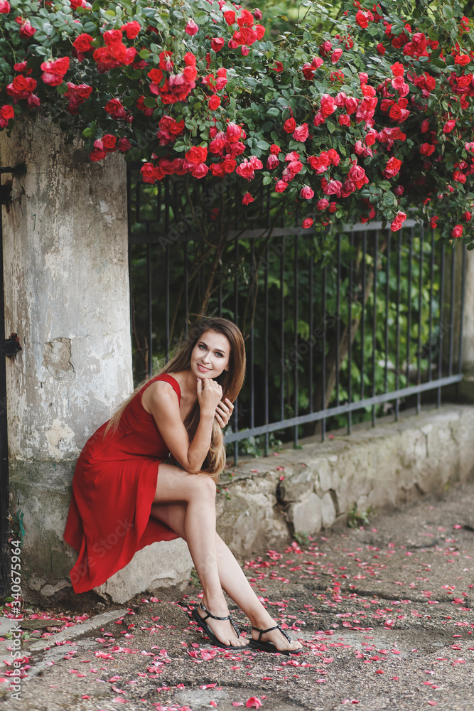 Beautiful young woman with long hair, wearing red dress, posing near flowering rose bushes on the old city street.