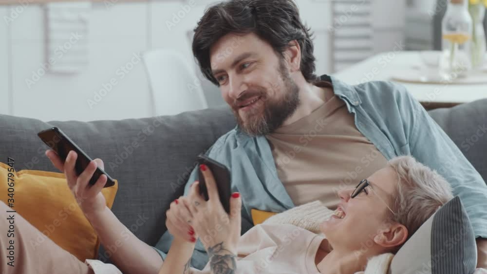 Young positive couple bonding on couch in the living room, browsing the Internet on their smartphones and chatting about something while staying together at home on quarantine Young positive couple bonding on couch in the living room, browsing the Internet on their smartphones and chatting about something while staying together at home on quarantine