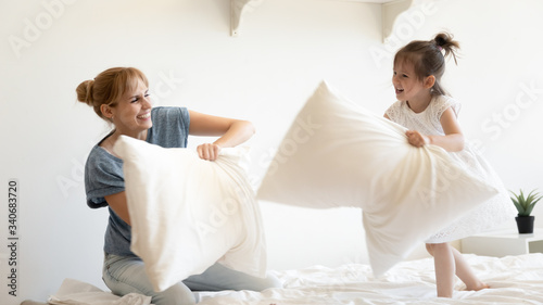 Happy young mother pillows fight and laughing with daughter on bed at bedroom. Smiling family of mom and little cute girl having fun and playing with cushions at home together.