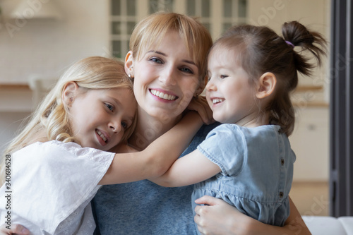 Close up headshot portrait picture of smiling mother hugging two daughter sitting on coach at home. Happy young parent with little children embracing looking at camera posing for photo in living room.