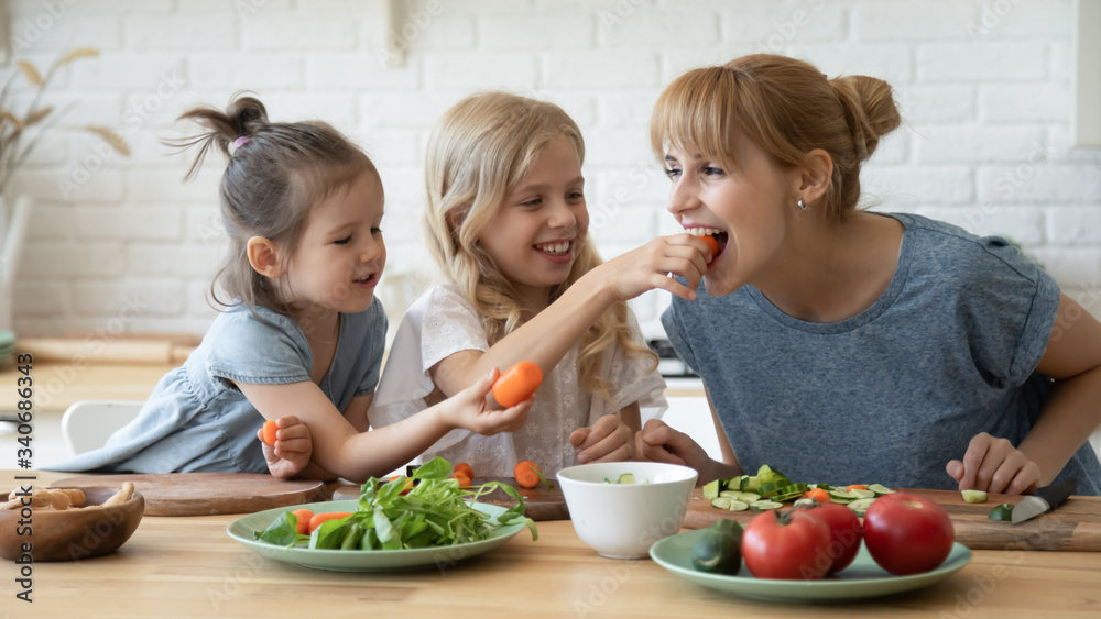 Caring daughter feeding mother teaching little children standing at table in kitchen. Happy mom with girls sisters chopping vegetables lettuce on wooden cutting board, preparing dinner.