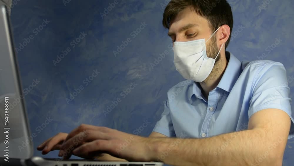 Remote work at home. Masked office worker, a man in a blue shirt sits at home and works. Types on a laptop Remote work at home. Masked office worker, a man in a blue shirt sits at home and works. Types on a laptop