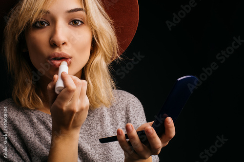 Beautiful amazing young woman using lipstick for lips. Putting some make up on face. Posing on camera. Hold fasial powder with small mirror in it. Isolated over black background. photo