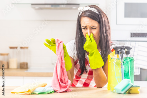 Female holding her nose with fingers upon smelly rag held in the other hand, leaning on kitchen desk, with cleaning stuff surrounding her photo