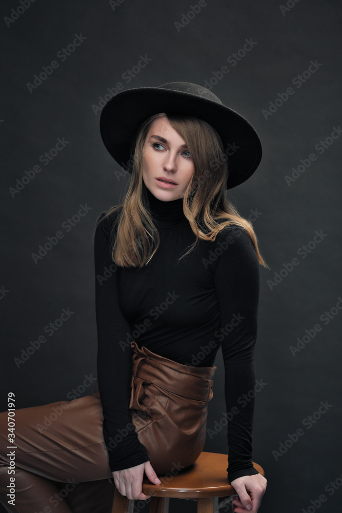 Portrait of young beautiful blond woman with light makeup and blue eyes touching her face. Dark background. Black tight sweater and hat with fields. Portrait of young beautiful blond woman with light makeup and blue eyes touching her face. Dark background. Black tight sweater and hat with fields.