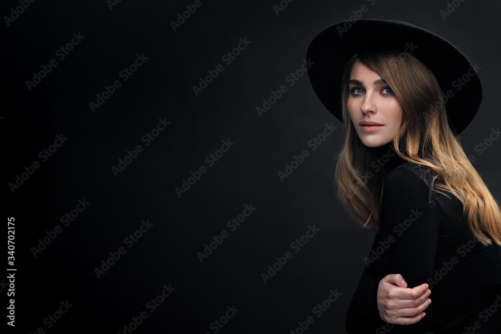 Portrait of young beautiful blond woman with light makeup and blue eyes touching her face. Dark background. Black tight sweater and hat with fields. Portrait of young beautiful blond woman with light makeup and blue eyes touching her face. Dark background. Black tight sweater and hat with fields.