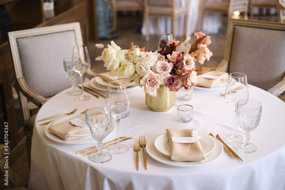 Fototapeta premium Luxury cozy autumn wedding table decoration in the restaurant. Fresh and dried flowers, roses, carnations. Beautiful table setting: golden appliances, fork and knife, calligraphy guest seating card.