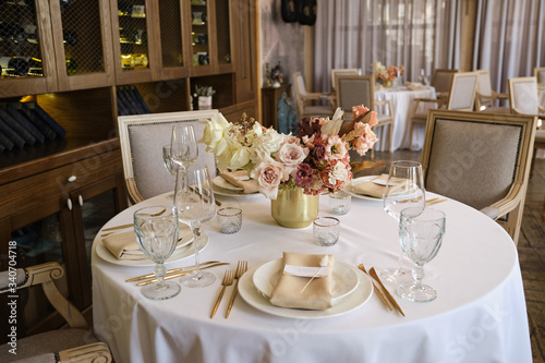 Luxury cozy autumn wedding table decoration in the restaurant. Fresh and dried flowers, roses, carnations. Beautiful table setting: golden appliances, fork and knife, calligraphy guest seating card.