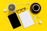Flat lay of modern home office workspace with tablet, coffee cup, note pad, pen, eyeglasses and cactus on yellow background. Top view