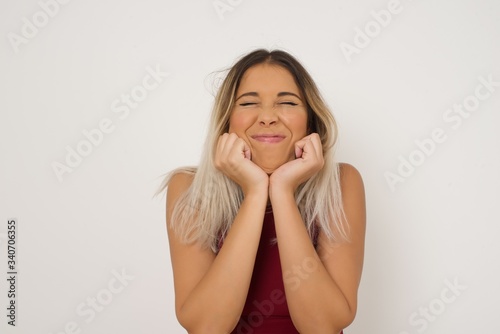 Portrait of young cute european student being overwhelmed with emotions, expressing excitement and happiness with closed eyes and hands near face while smiling broadly over gray background.
