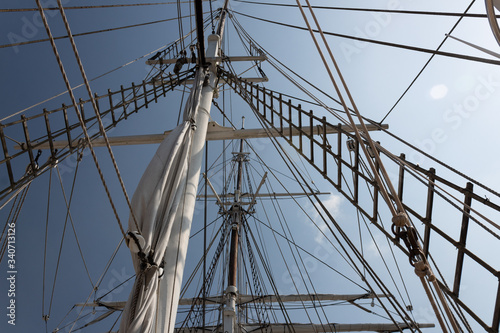 Two masts of an old tall ship, rigging and shrouds seen from below, blue sky and sun, horizontal aspect