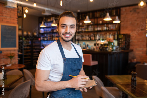 Happy young waiter in white t-shirt and dark blue apron looking at you