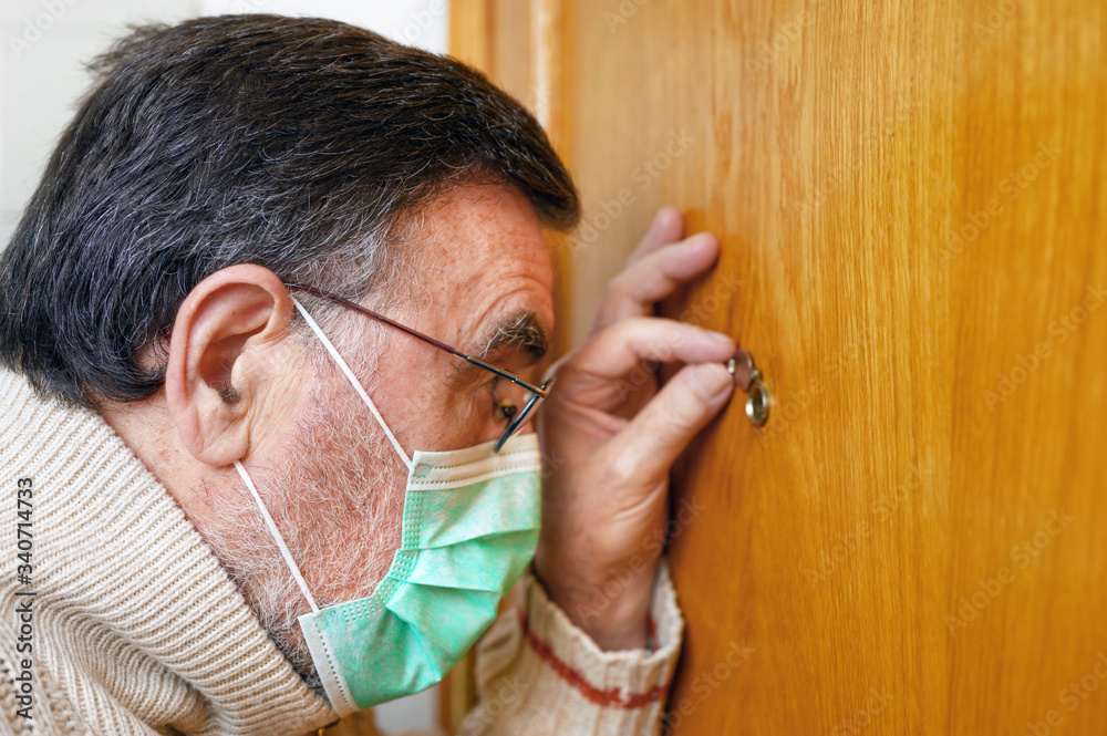 Obraz premium Covid-19 concept. Stay at home. Self-isolation to prevent the coronavirus pandemic. Senior man in protective green mask looks through the peephole .