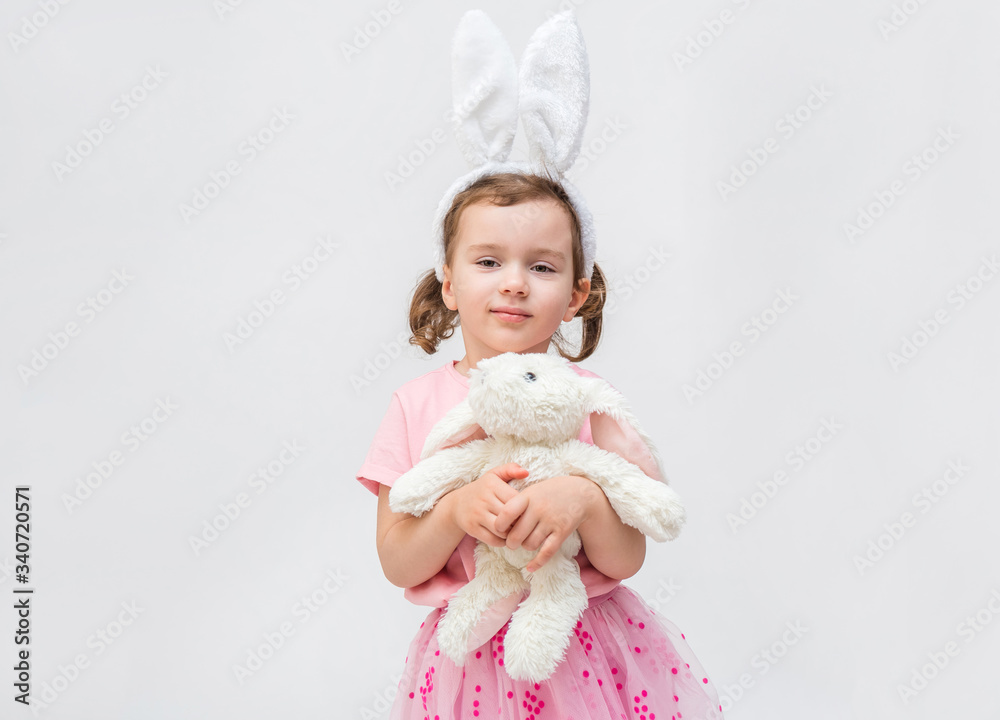A cute girl on a white background with an empty seat. A girl with hare ears and a stuffed rabbit toy. A little girl in a pink t-shirt. The girl looks at the camera