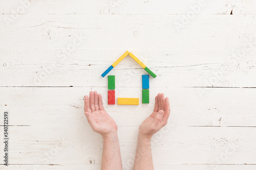 Directly above on wooden colorful bricks on a white wooden background. Hands of a man protect a house assembled from multi-colored wooden blocks. Home, family and education concept.