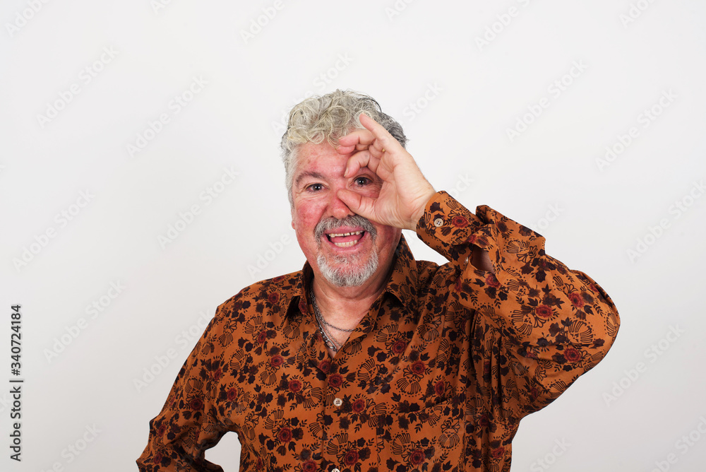 Gorgeous young blond man with happy face smiling doing ok sign with hand on eye looking through finger. Wearing casual clothes and standing. Gorgeous young blond man with happy face smiling doing ok sign with hand on eye looking through finger. Wearing casual clothes and standing.