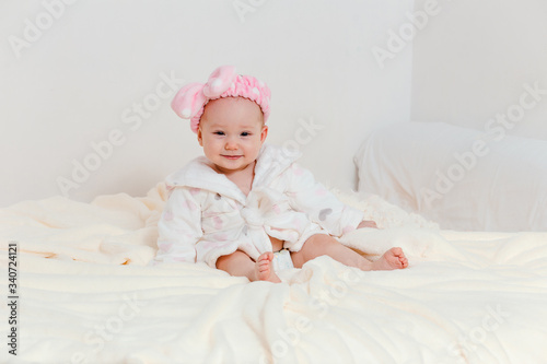 laughing baby girl in a Terry robe and diaper sitting on the bed after bathing. an eight month old baby girl with a pink headband