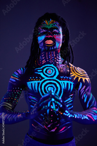 portrait of man having fluorescent make-up and body art glowing on neon lights, african guy stand in the pose of prayer