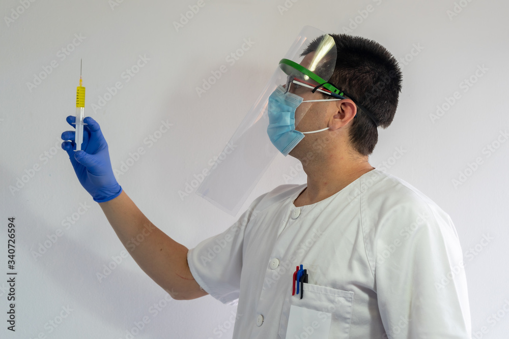 Naklejka premium Doctor / Nurse with face shield observes the coronavirus vaccine. In his hand he has the vaccine and a syringe. He wears a white uniform, wears a mask, gloves and glasses.