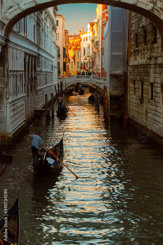 Several gondolas sail in a row along the narrow venetian canal in the light of the setting sun. © karinabost