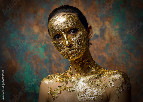 Girl with a mask on her face made of gold leaf. Gloomy studio portrait of a brunette on an abstract background. © Mountains Hunter