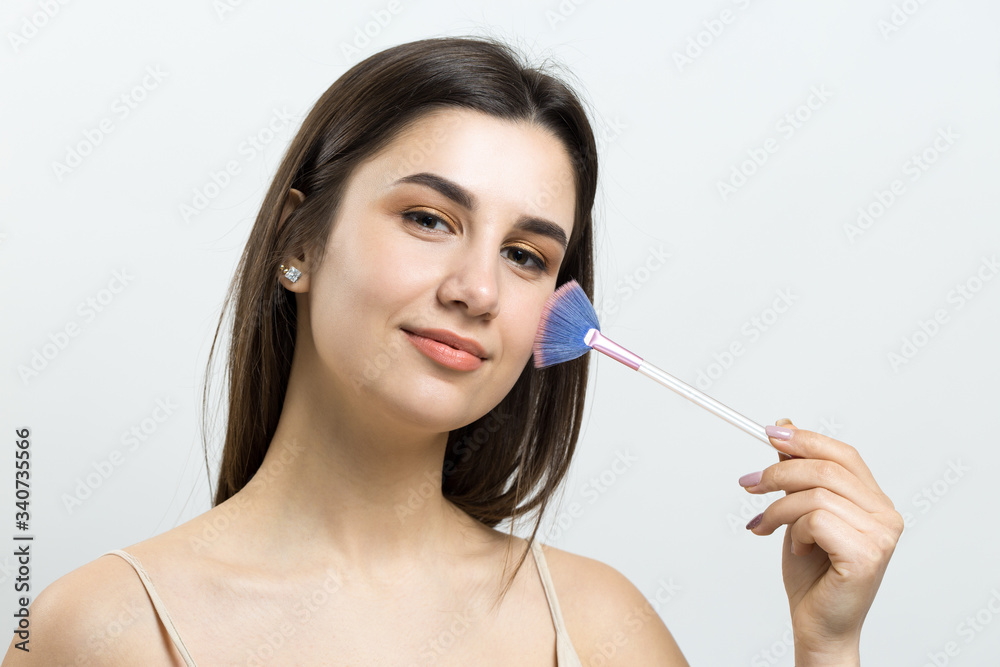 Close-up of a young girl in a light top on a white background making a facial make-up. A pretty woman holds a cosmetic brush near her face and smiles. Close-up of a young girl in a light top on a white background making a facial make-up. A pretty woman holds a cosmetic brush near her face and smiles.