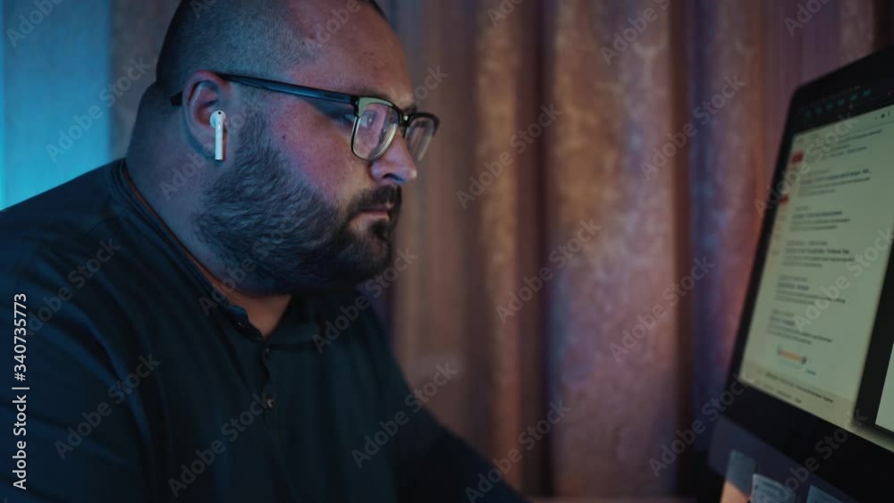 Distant work, man in glasses working at computer and talking by earphones at night, freelancer at home Distant work, man in glasses working at computer and talking by earphones at night, freelancer at home