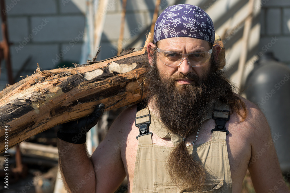 A middle-aged bearded man in a bandana cuts logs with an ax. Brutal in overalls does the hard work on a hot day.