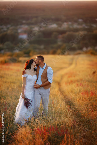 Young beautiful stylish couple in a flowering field of wheat on a road on a hill with a view of the city at sunset. Rustic wedding photo shoot. Bride and groom in a white dress with a train © Саша Тихова