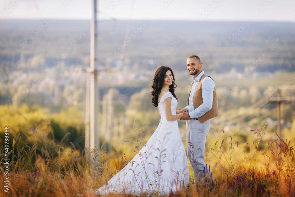 Happy beautiful couple in love hold each other hands in flowers on a field on a hill. Stylish wedding photo session in rustic style. Romantic portrait of the bride and groom on their wedding day