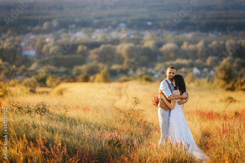 Young beautiful couple in a flowering field on a hill hug each other at sunset. Rustic wedding photoshooting. Romantic sensual portrait of the bride and groom on their wedding day