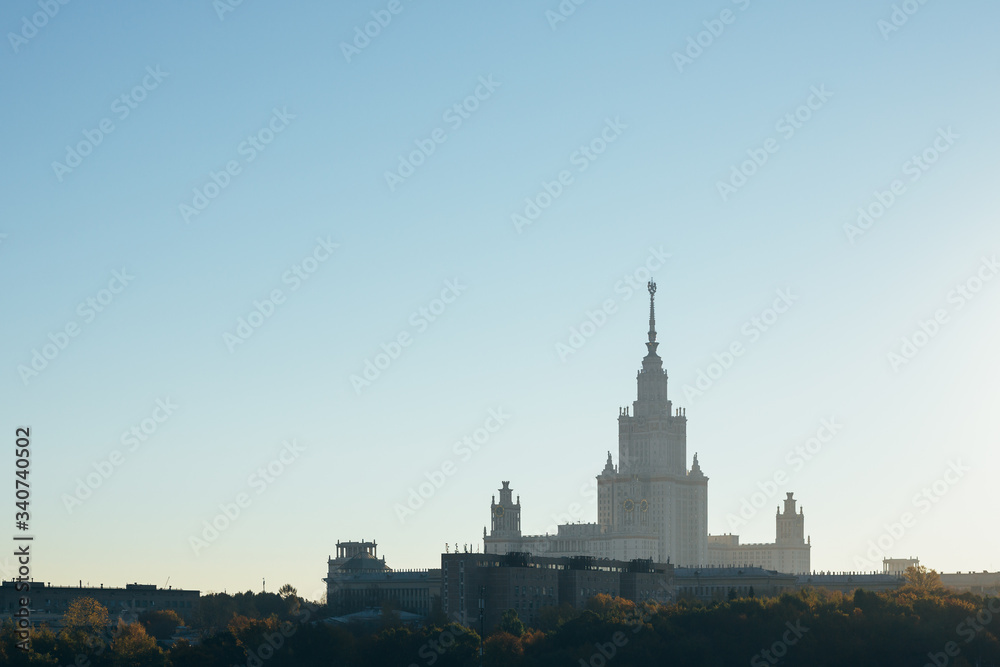 Fototapeta premium Moscow landscape. Moscow University building in morning haze behind autumn colored trees and smaller buildings. There is lot of negative space in clear sky with the sunrise glow on the right side.