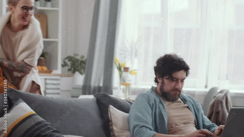 Bearded Caucasian man sitting on couch and working on laptop at home. Joyous wife hitting him with pillow and laughing, then playful couple having pillow fight in the living room Bearded Caucasian man sitting on couch and working on laptop at home. Joyous wife hitting him with pillow and laughing, then playful couple having pillow fight in the living room
