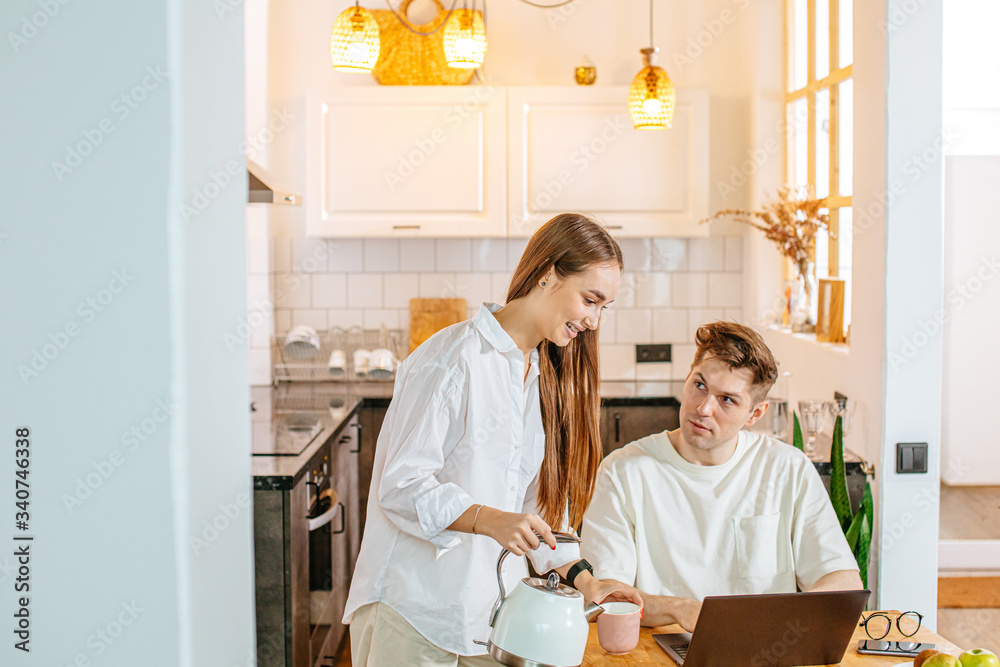 attractive careful lady pouring tea from kettle to give it for working husband. kind wife take care of her working husband, he use laptop, freelance attractive careful lady pouring tea from kettle to give it for working husband. kind wife take care of her working husband, he use laptop, freelance
