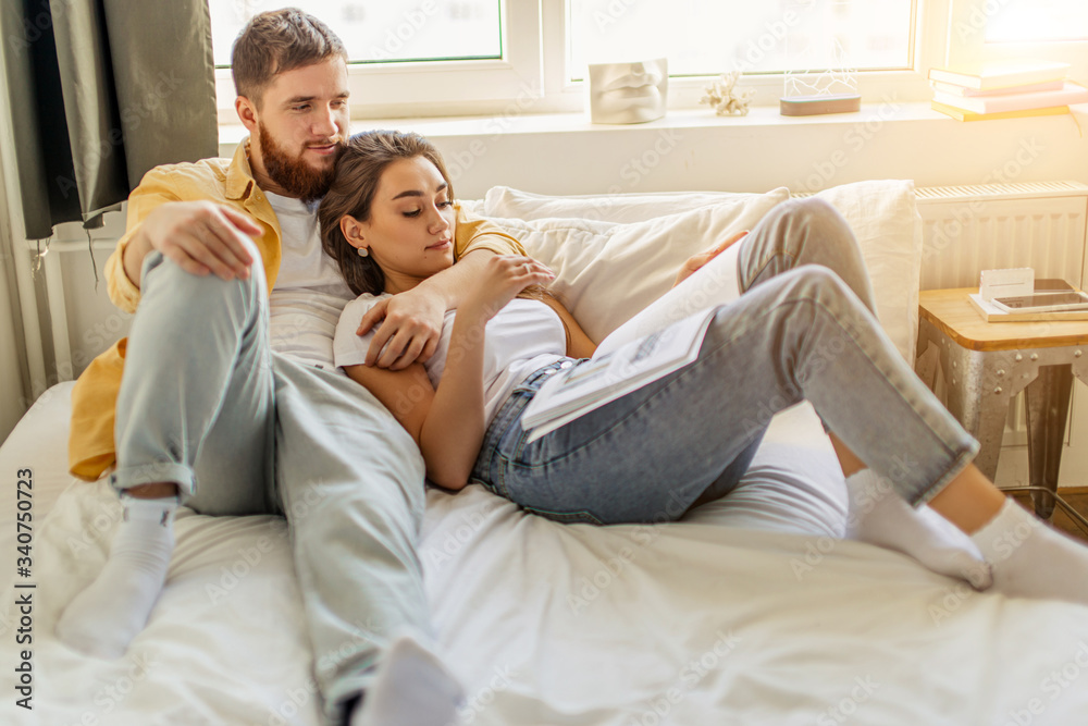 weekends, holidays of young married caucasian couple at home. happy man and woman spend time together, lie on bed and read book. weekends, leisure, free time indoors