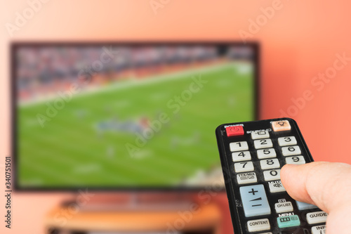 Caucasian man watching american football game on TV at home. Changing channels and adjusting volume with television remote control. 