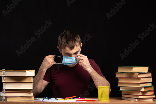 man puts on a medical mask while sitting at a table with books in the office. Black background. quarantined remote work
