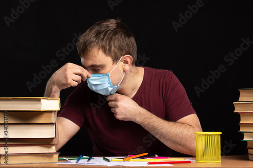 a young man sits at a desk with books and adjusts a medical mask on his face. quarantine.