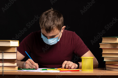 a young man in a medical mask sits at a table surrounded by books and writes something. quarantined work. Black background. isolate