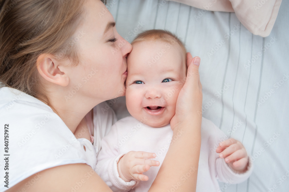 Mother kissing her infant baby in bed. Happy mother kisses baby lying on the bed. Beautiful smiling girl lying on her back. Top view. Flat Lay. Mother kissing her infant baby in bed. Happy mother kisses baby lying on the bed. Beautiful smiling girl lying on her back. Top view. Flat Lay.